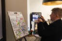 The artist Matthew Buck from Drawnalism takes a photo of his sketch of Taylor Morrison's Self Care presentation. The sketch is a series of cartoon scenes depicting the various points that Taylor made. The sketch is made on an A, 2 sketch pad positioned on an easel in the corner of the conference room.
