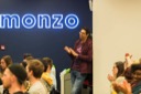 A smiling Akash Goswami stands at the back of the conference room and applauds the end of Sam Warner's presentation. The blue neon Monzo sign dominates the background.