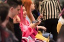 Rachel Konichiwakitty sits in the audience and looks up from her phone. She is wearing a pink cardigan adorned with dark pink hearts. The cardigan matches her pink hair.