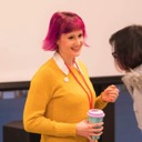 Jo Franchetti smiles as she talks with a conference attendee. She is holding a re-usable coffee cup and listening to the person speak. Jo is wearing a yellow sweater and she has bright pink hair styled in a bob.