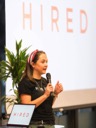 Libby Derbyshire from Hired speaks to the conference. She stands in front of a large screen featuring the Hired logo. Her long hair is tied back with a red and white striped silk scarf and she wears a black t-shirt with Hired written in white uppercase letters on the front.