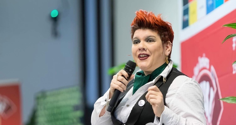 A close-up of a smiling Scary Boots delivering their conference opening. They wear a white shirt with a dark green cravat and a black waistcoat. Their short styled hair is a fiery red and they wear a silver nose ring.