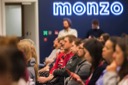 A view along a row of the audience as they listen to Sascha Wolf's speech. In the background the Monzo logo is emblazoned across the wall in blue neon. The room is crowded and audience members stand at the back, beneath the Monzo sign.