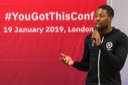 A close-up of Nathaniel Okenwa, from Twilio, as he speaks at the You Got This Conference. He wears a black hoodie with a white zip and is emphasizing his point by gesturing with his right hand.