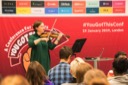 Paula Muldoon, a JavaScript Developer at Cambridge Cognition, opens her session entitled, Where Did All My Money Go?, by playing the violin. She stands at the front of the conference room against a backdrop of posters featuring You Got This sponsors.