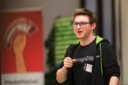 Tim Fogarty, a software engineer at Mongo D, B, smiles as he takes to the stage. He holds a microphone in his right hand and he wears a black Mongo D, B t-shirt and a black and lime-green hoodie. Tim wears glasses and cuts an energetic figure.