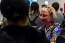 A conference attendee, wearing a rainbow coloured shirt and heart-shaped earrings, smiles as she chats with other attendees in the conference foyer. Her blonde hair is held in place with small, brightly coloured hair clips.