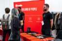 A conference attendee talks to a smiling Sam Bellem from Auth 0 at his stand. In the background, Nathaniel Okenwa is visible at the Twilio stand.