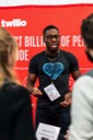 Nathaniel Okenwa talks to a couple of conference attendees at the Twilio stand. He is wearing a dark grey t-shirt featuring a blue heart motif containing the words, Sendgrid loves start-ups.