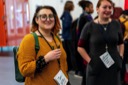 Amy Dickens, one of the conference organisers, smiles for the camera as they stand in the conference foyer. They are wearing a mustard coloured jumper, large, dark-framed glasses and a green backpack.