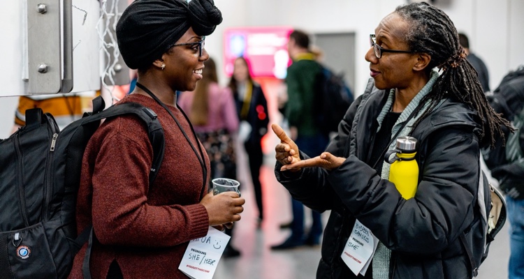 A pair of conference attendees are deep in conversation in the foyer. The woman on the left, wearing a black head scarf, is smiling broadly as she listens to her companion, who has flowing braids.
